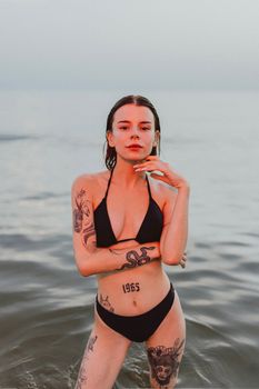 Woman with tattoos standing confidently in a bikini at the ocean shore.