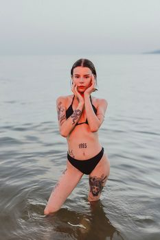 A woman with tattoos stands confidently in the sea wearing a bikini, capturing an artistic beach portrait.