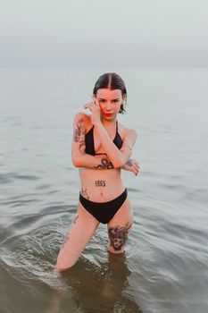 A tattooed woman in a black bikini stands confidently in the ocean, exuding style and attitude.
