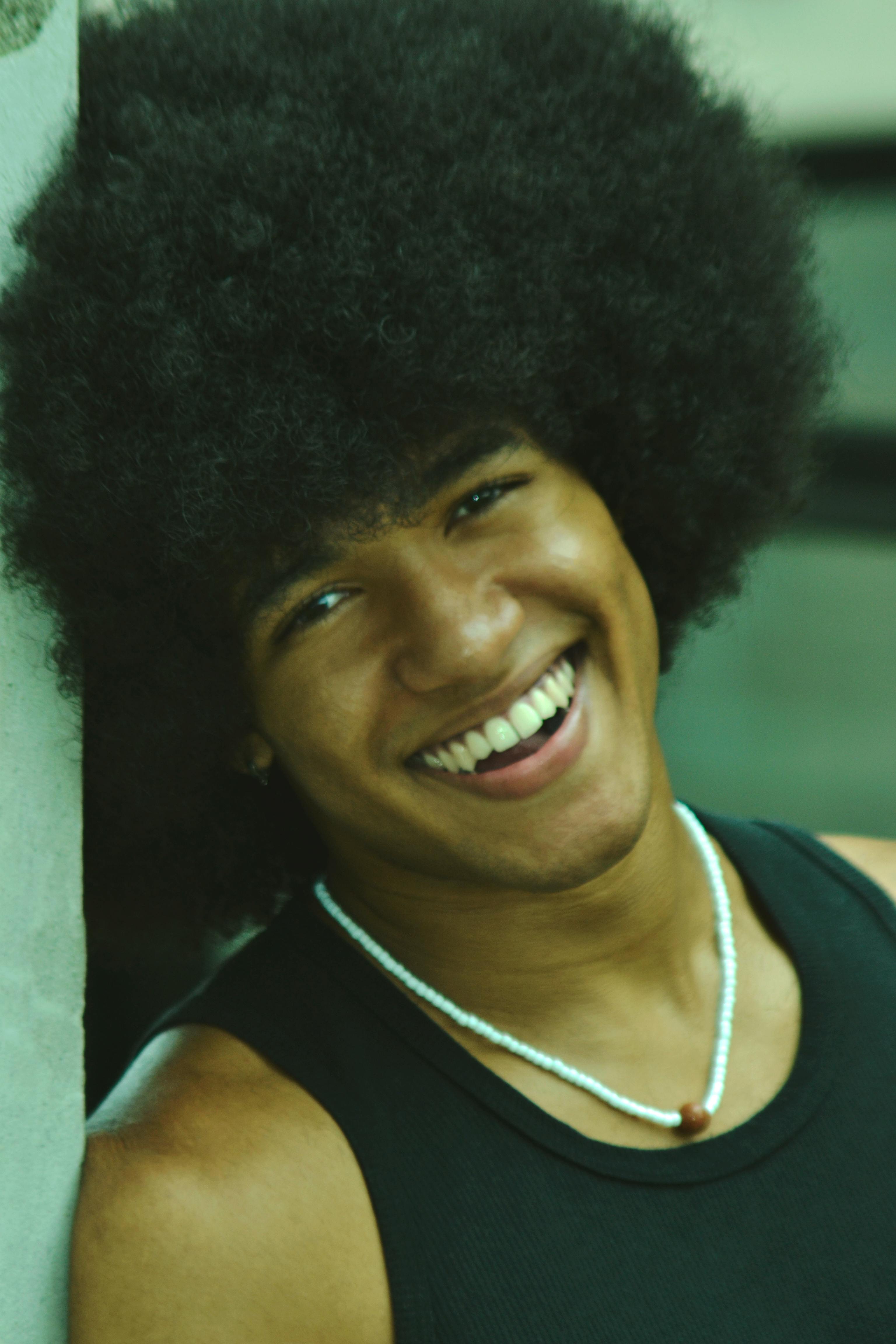 A young man with an afro hairstyle smiles brightly outdoors, showcasing happiness and style.