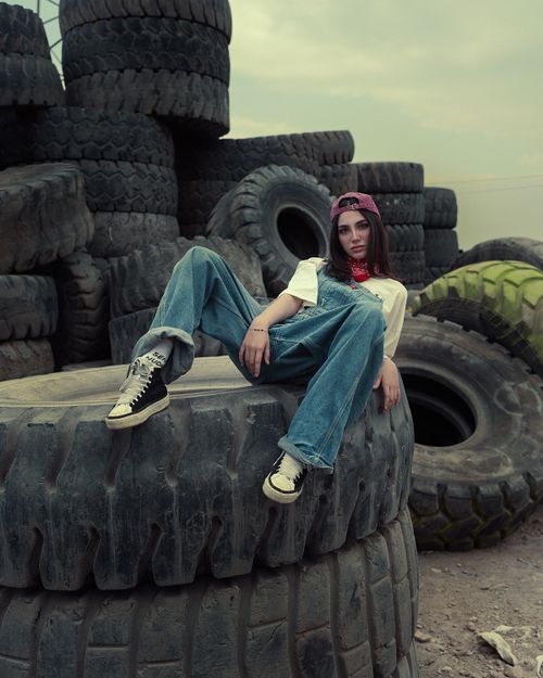 Free Stylish young woman in jeans and cap poses on large stacked tires outdoors. Stock Photo