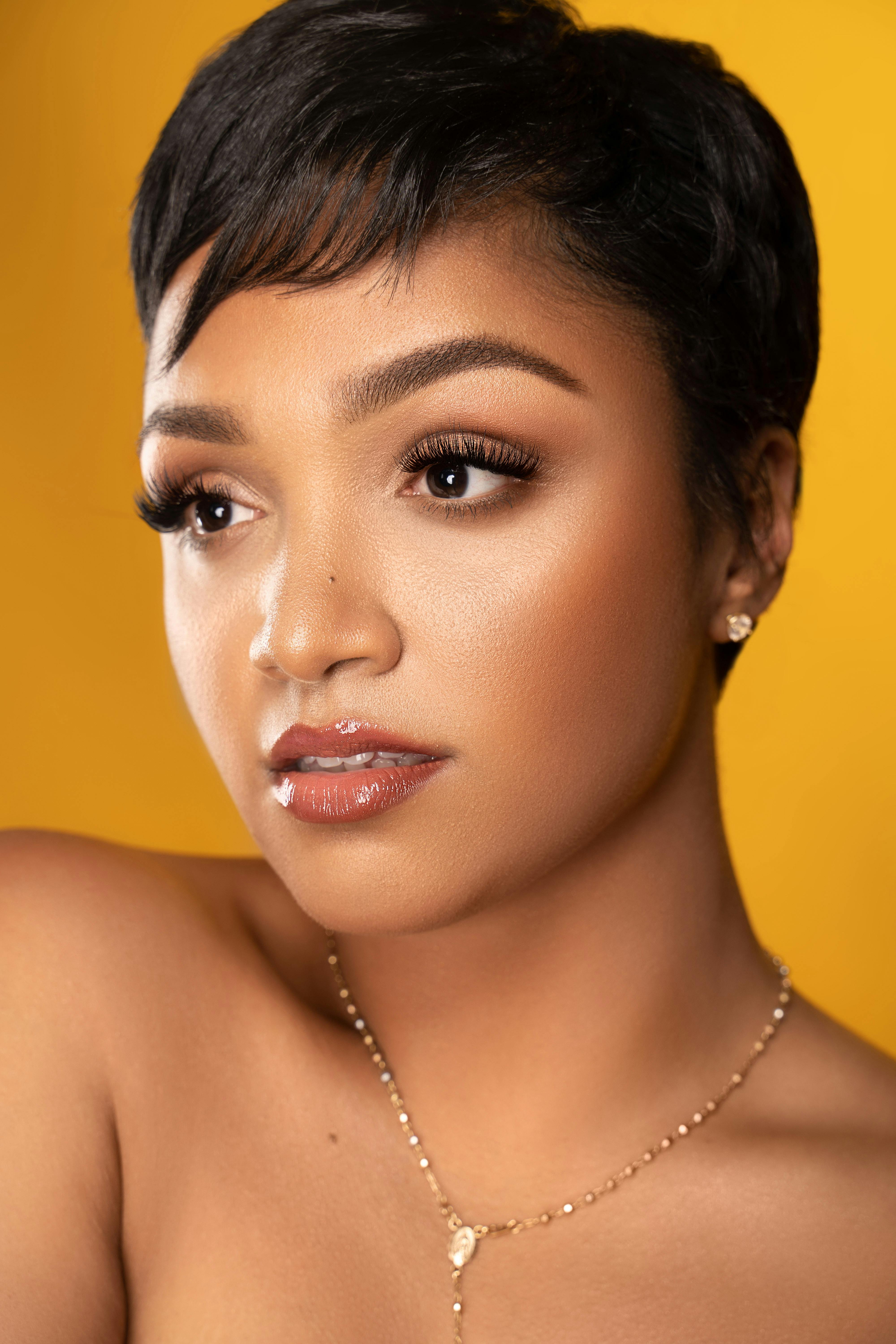 Close-up portrait of a stylish woman with short hair, makeup, and a necklace against a yellow background.