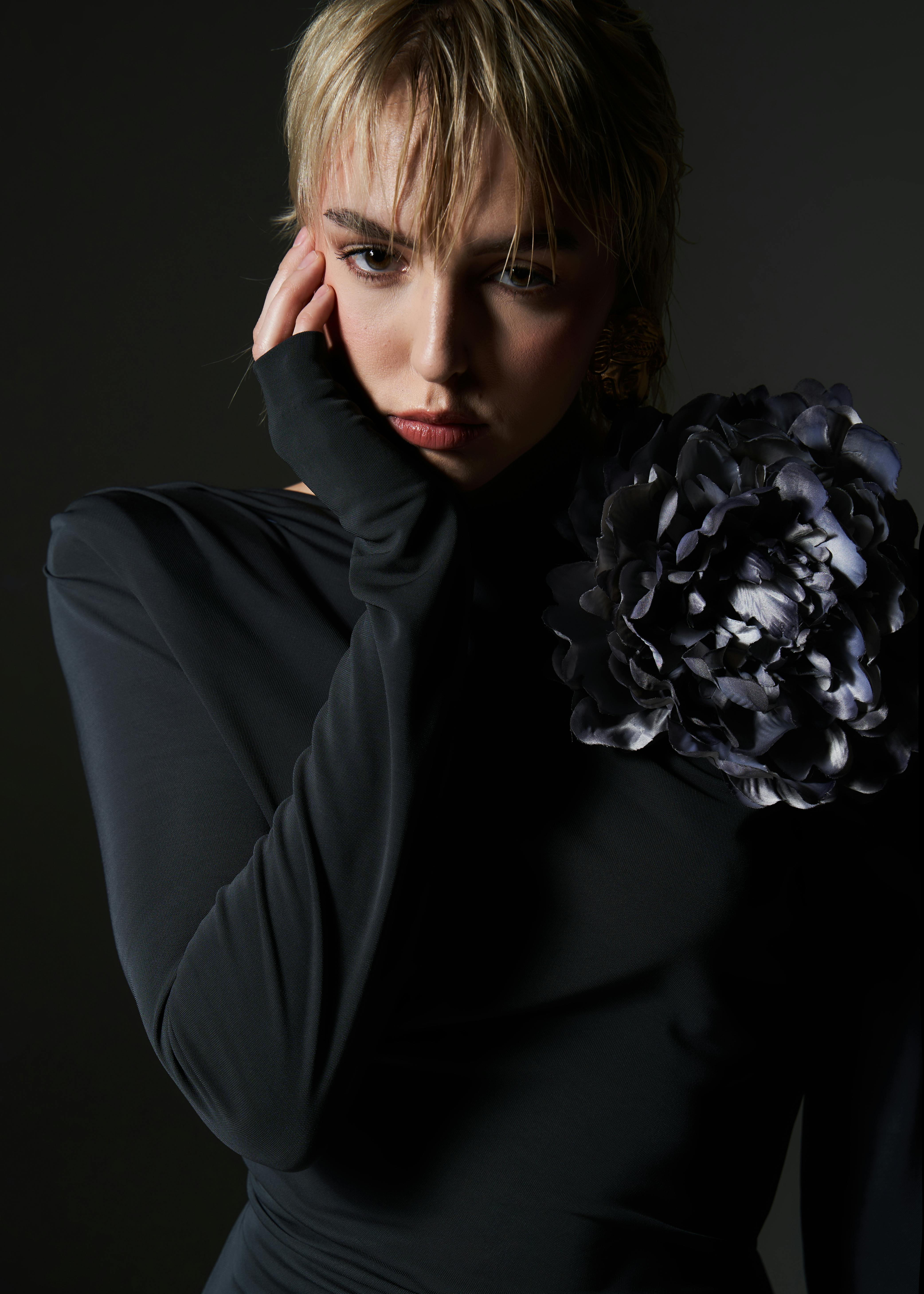 A stylish young woman poses in a dark studio, wearing a black dress adorned with a large floral design.