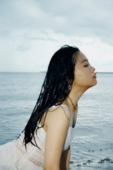 A woman with wet hair in a white dress enjoys the ocean breeze with eyes closed.