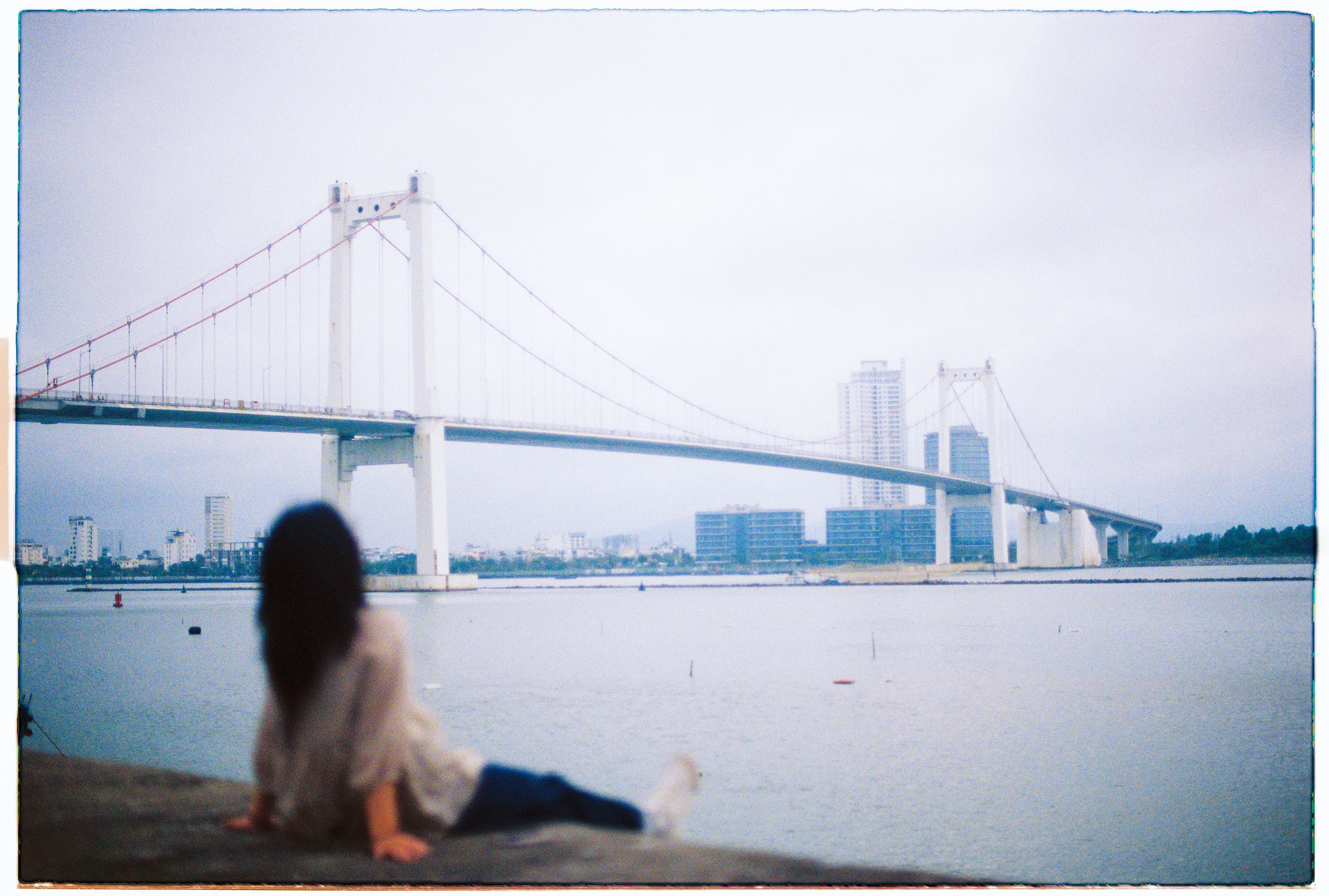 A woman sitting by the Han River with Thuan Phuoc Bridge and Da Nang skyline in the background.