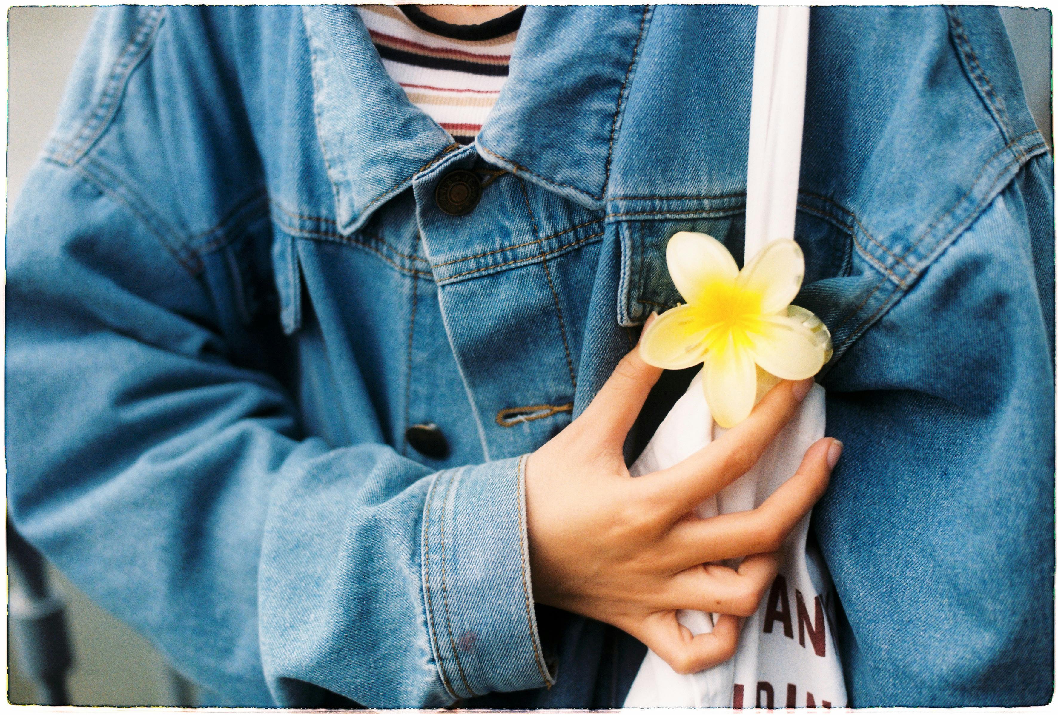 A person wearing a denim jacket holds a vibrant yellow plumeria flower against a tote bag.