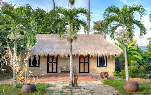 Free Tropical cottage with a thatched roof surrounded by palm trees in a lush garden. Stock Photo
