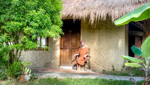 Free Asian woman sitting in a chair outside a rustic thatched-roof house, surrounded by lush greenery. Stock Photo