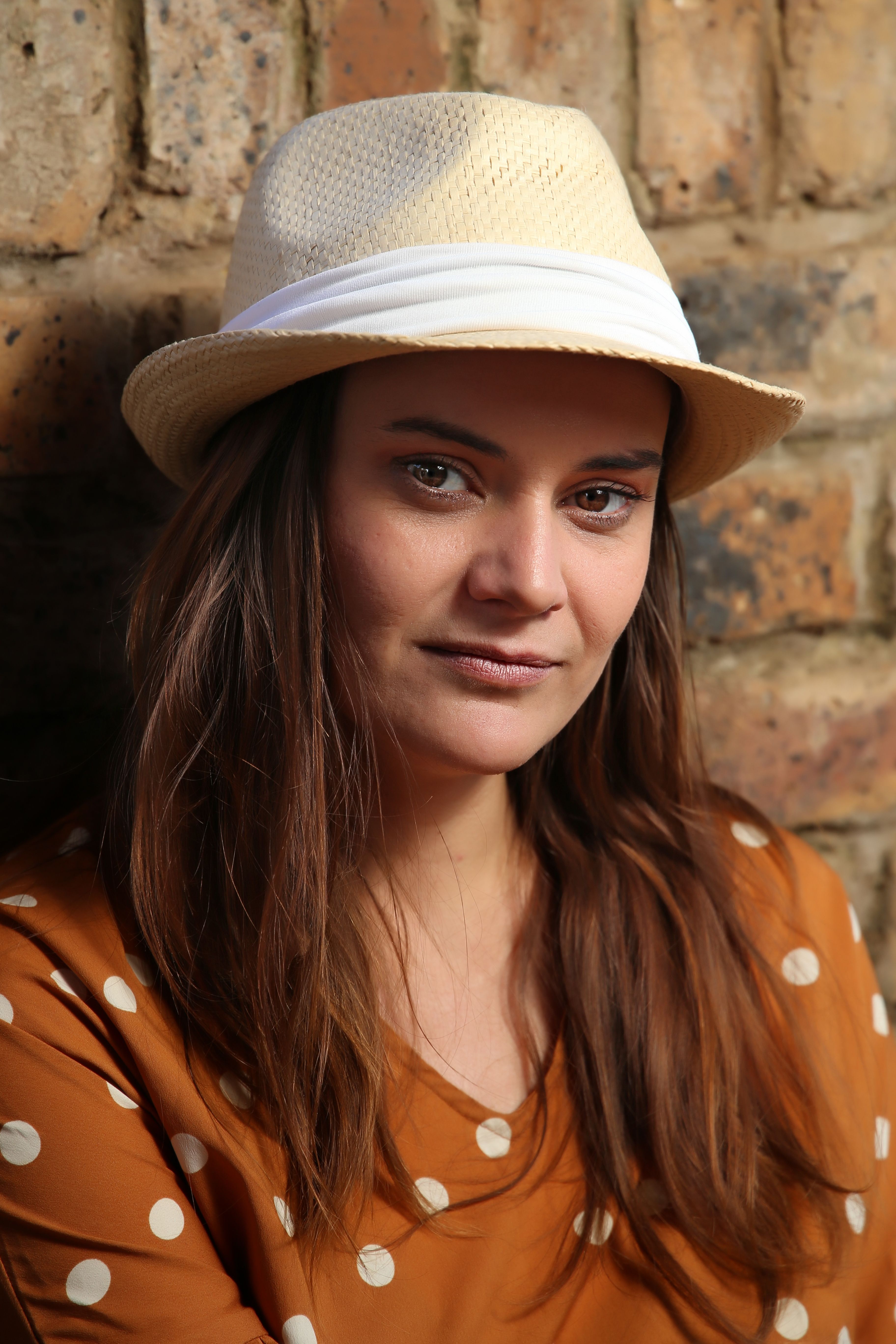 Stylish woman in a straw hat and polka dot dress posing against a brick wall.