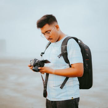 A young man standing outdoors, examining a camera with focus and interest.