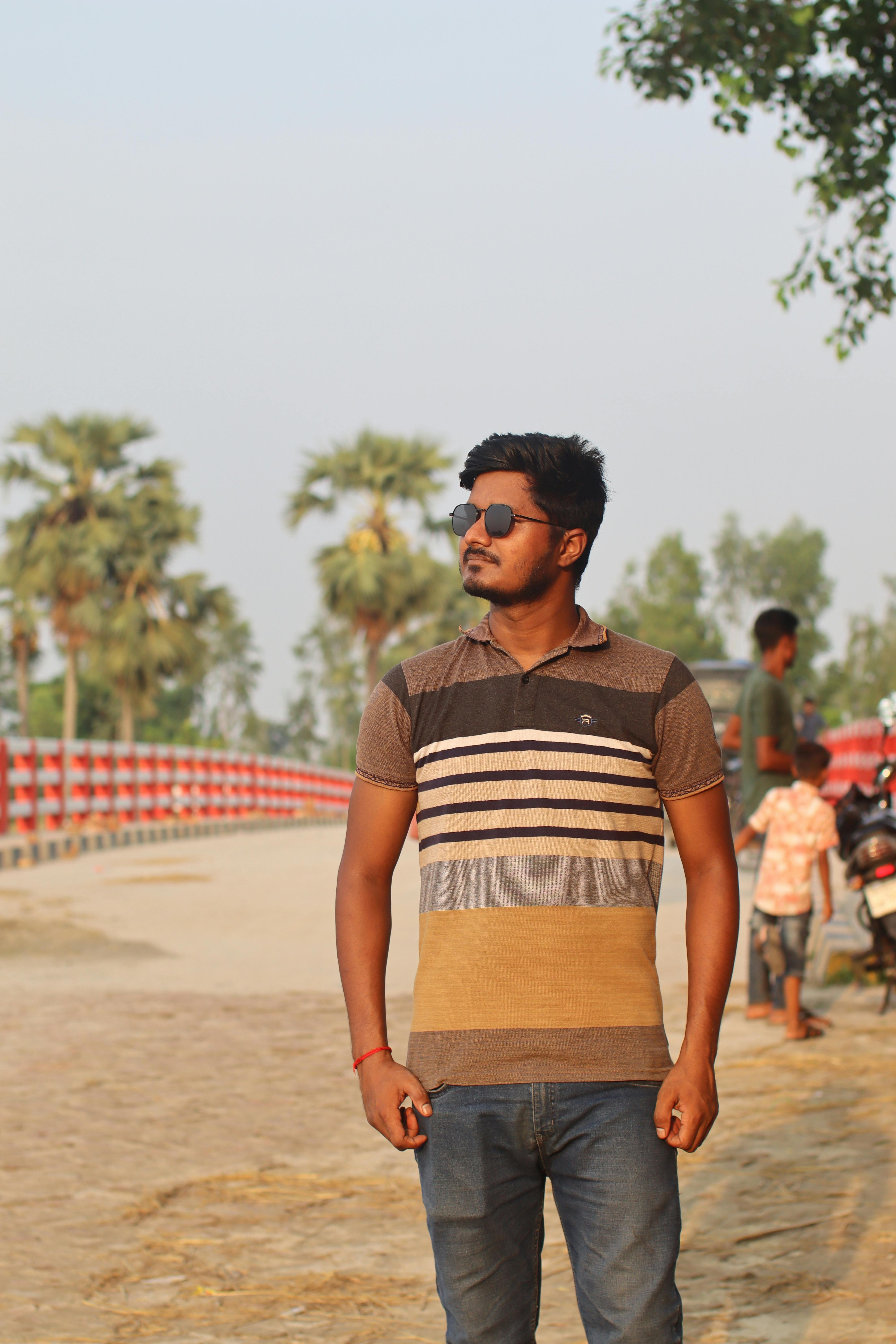 Young man with sunglasses poses stylishly in a rural area during the day.