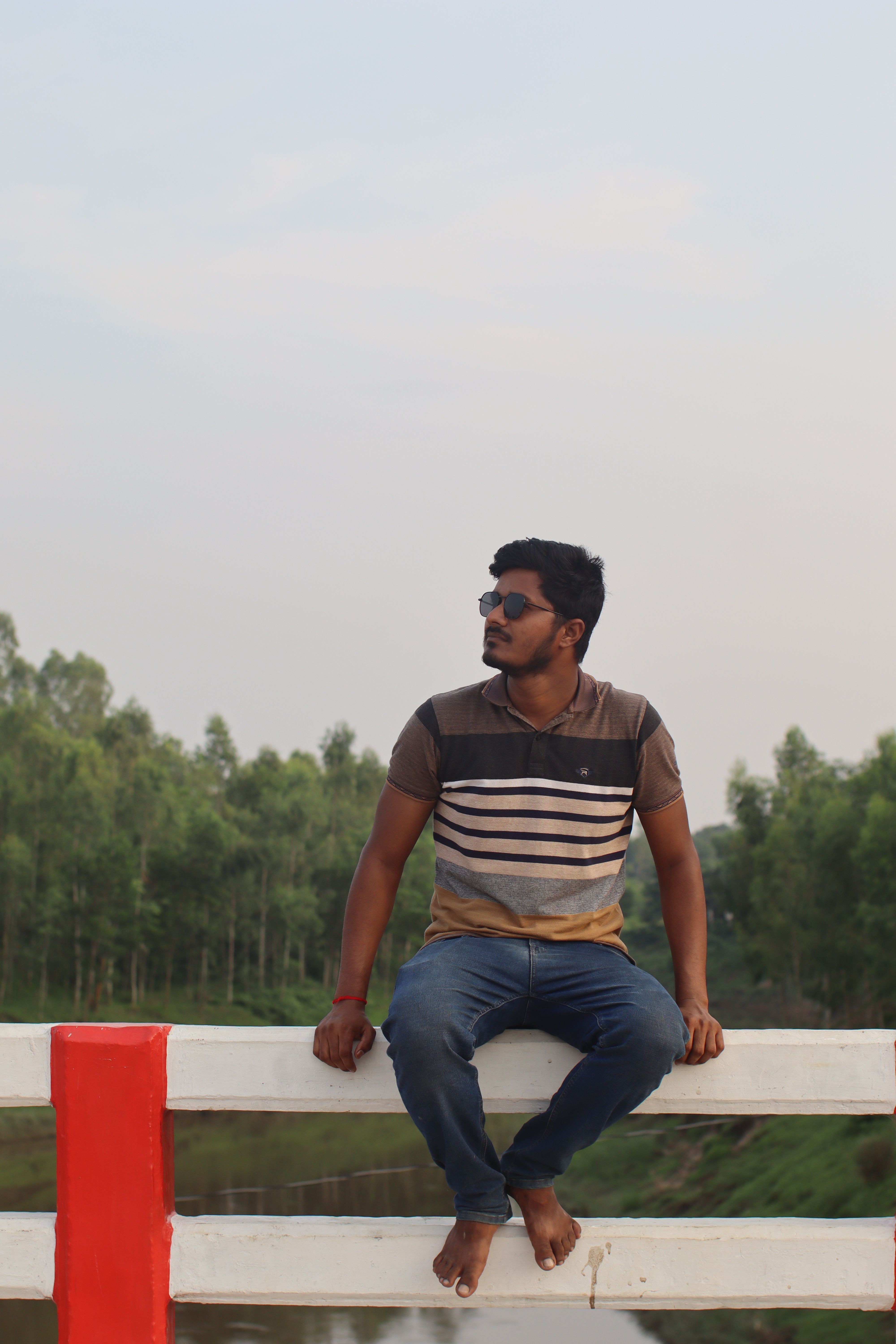 A young man in jeans and polo shirt sits barefoot on a fence with a scenic backdrop in Bangladesh.