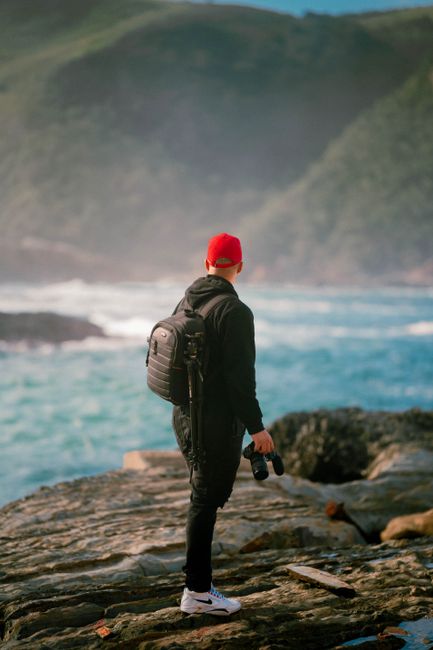 A man with a backpack and camera stands on a rocky shore in South Africa.