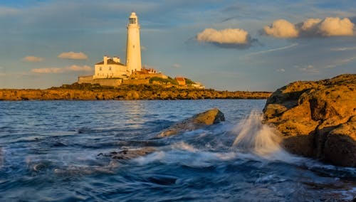 A lighthouse is seen in the distance with waves crashing on the rocks