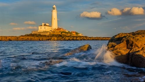 Free Stunning view of St Mary's Lighthouse on a rocky seashore with ocean waves under a vibrant sunset sky. Stock Photo