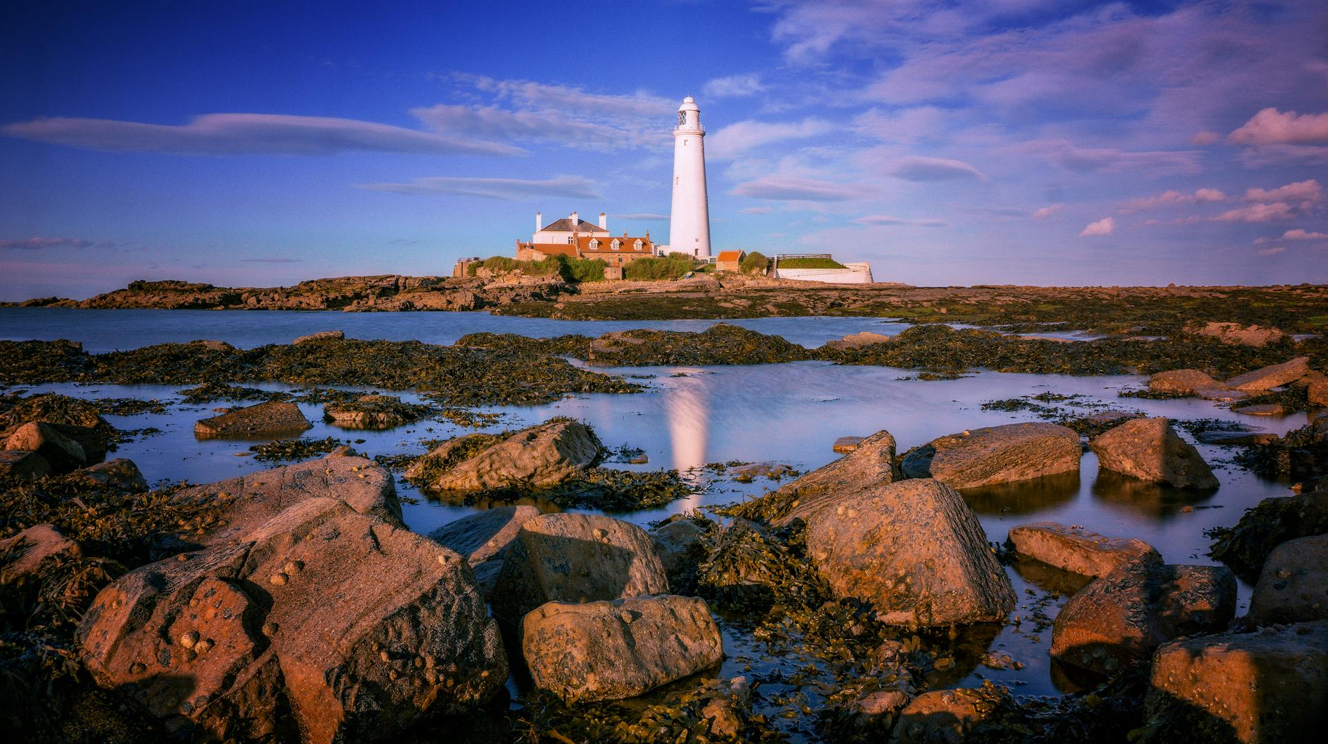 A picturesque lighthouse on a rocky coast reflected in serene waters under a vibrant sky.