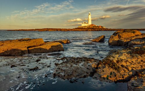 Free Scenic view of St Mary's Lighthouse surrounded by rocky shoreline during sunset, Northumberland, UK. Stock Photo