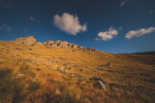 Free A stunning view of golden grass plains and rocky hills under a vibrant blue sky with white clouds. Stock Photo