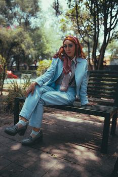 Fashionable woman in blue suit and headscarf sitting on a park bench, surrounded by trees.