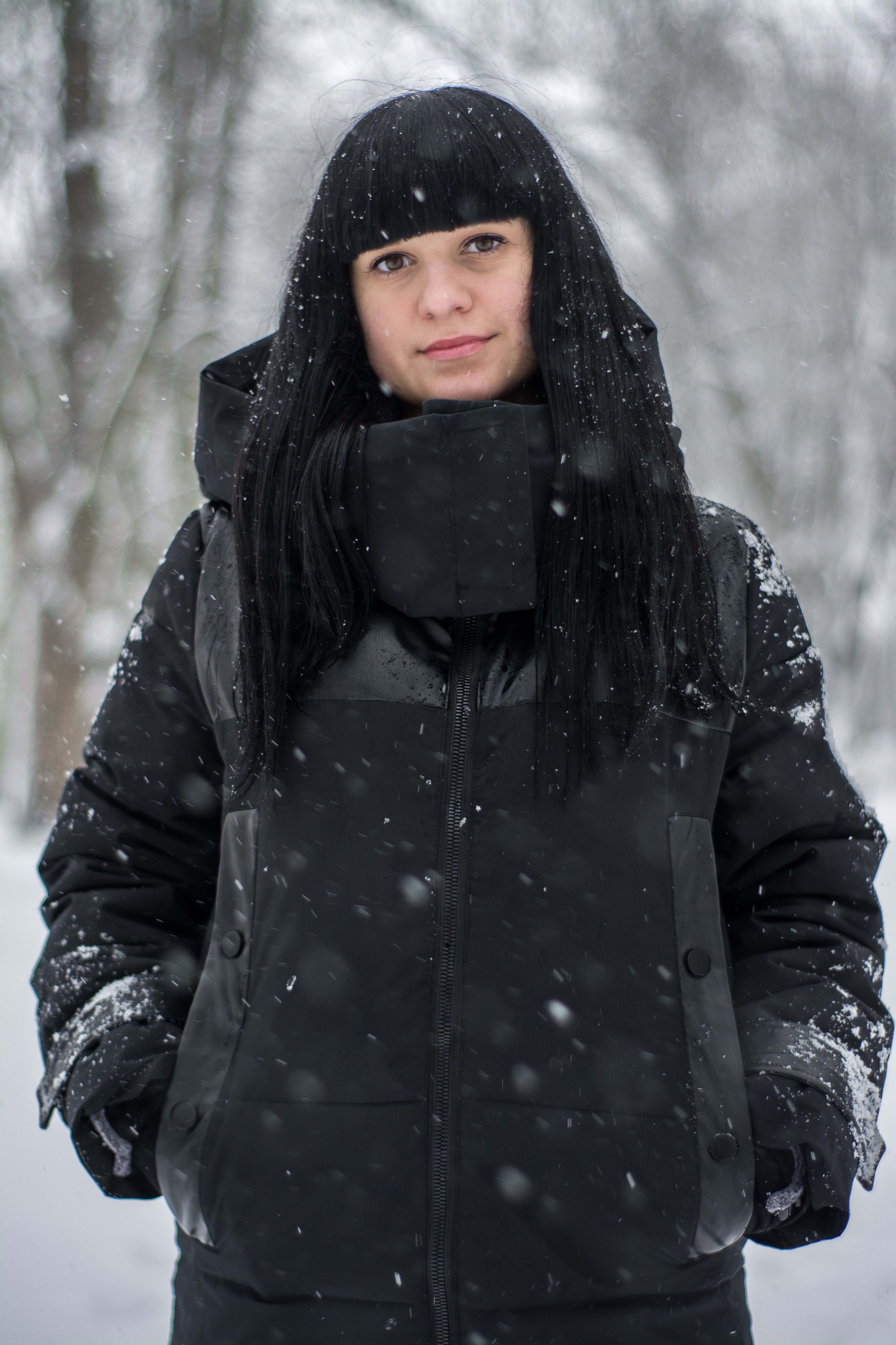 Young woman with bangs in a black coat standing in a snowy forest during winter.
