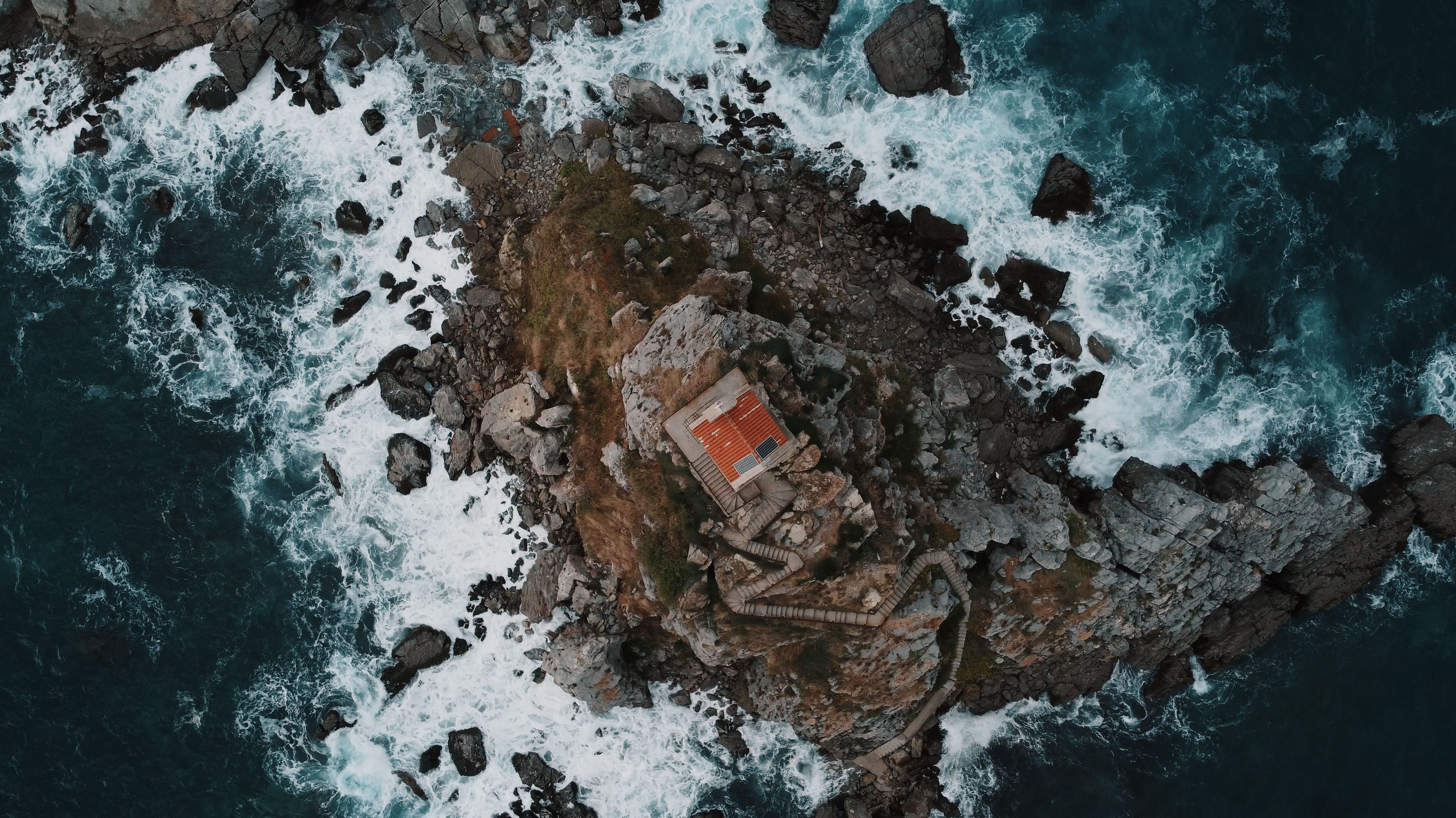 Drone shot of a church on a rocky coast in Petrovac, Montenegro. Waves crash against the shore in a dramatic display.