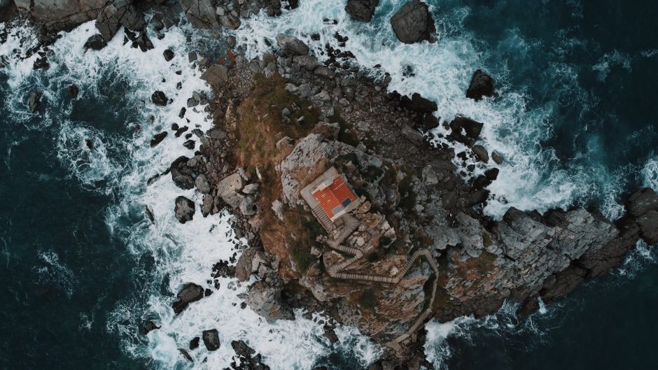 Drone shot of a church on a rocky coast in Petrovac, Montenegro. Waves crash against the shore in a dramatic display.