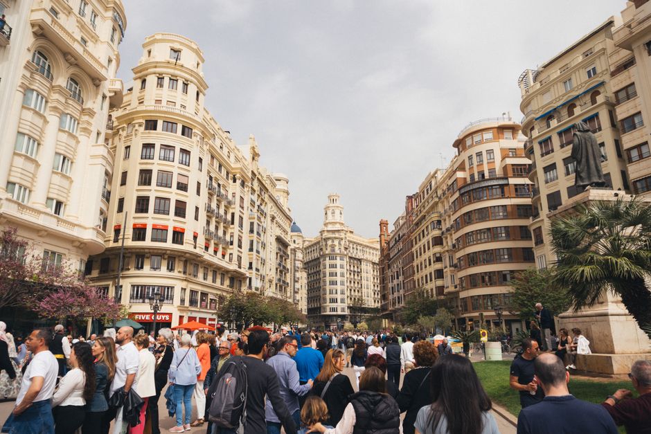 Una plaza bulliciosa en Valencia, España, con edificios históricos y una multitud diversa disfrutando de un día soleado.