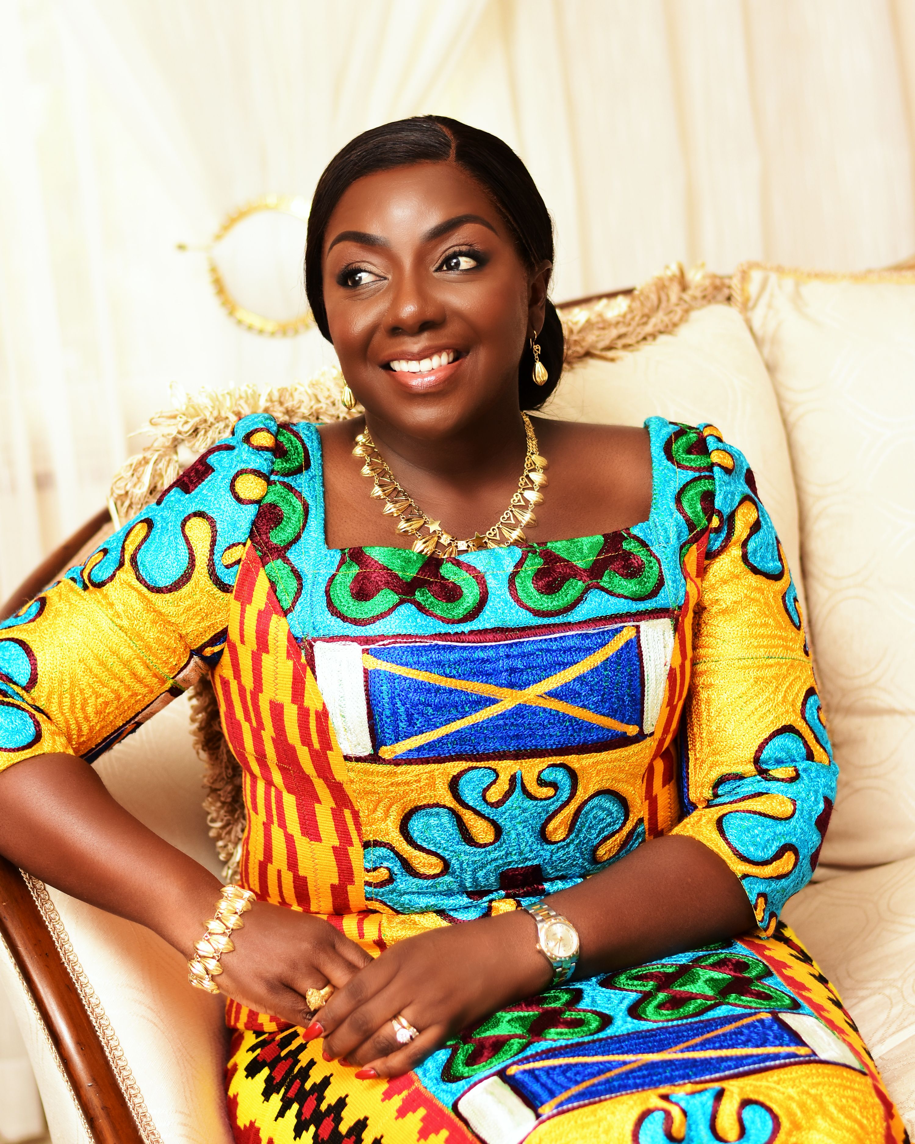 A joyful African woman in colorful traditional attire sitting indoors in Kumasi, Ghana.