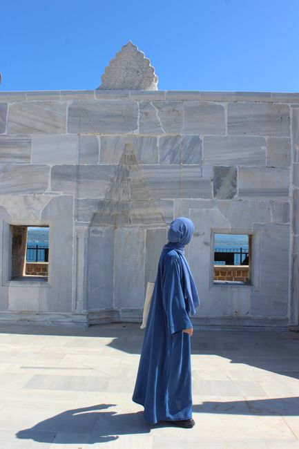 A woman in a blue hijab walks near an Ottoman mosque wall under a clear sky, Çanakkale, Turkey.