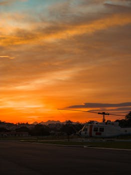 A helicopter sits on a helipad under a stunning orange sunset sky.