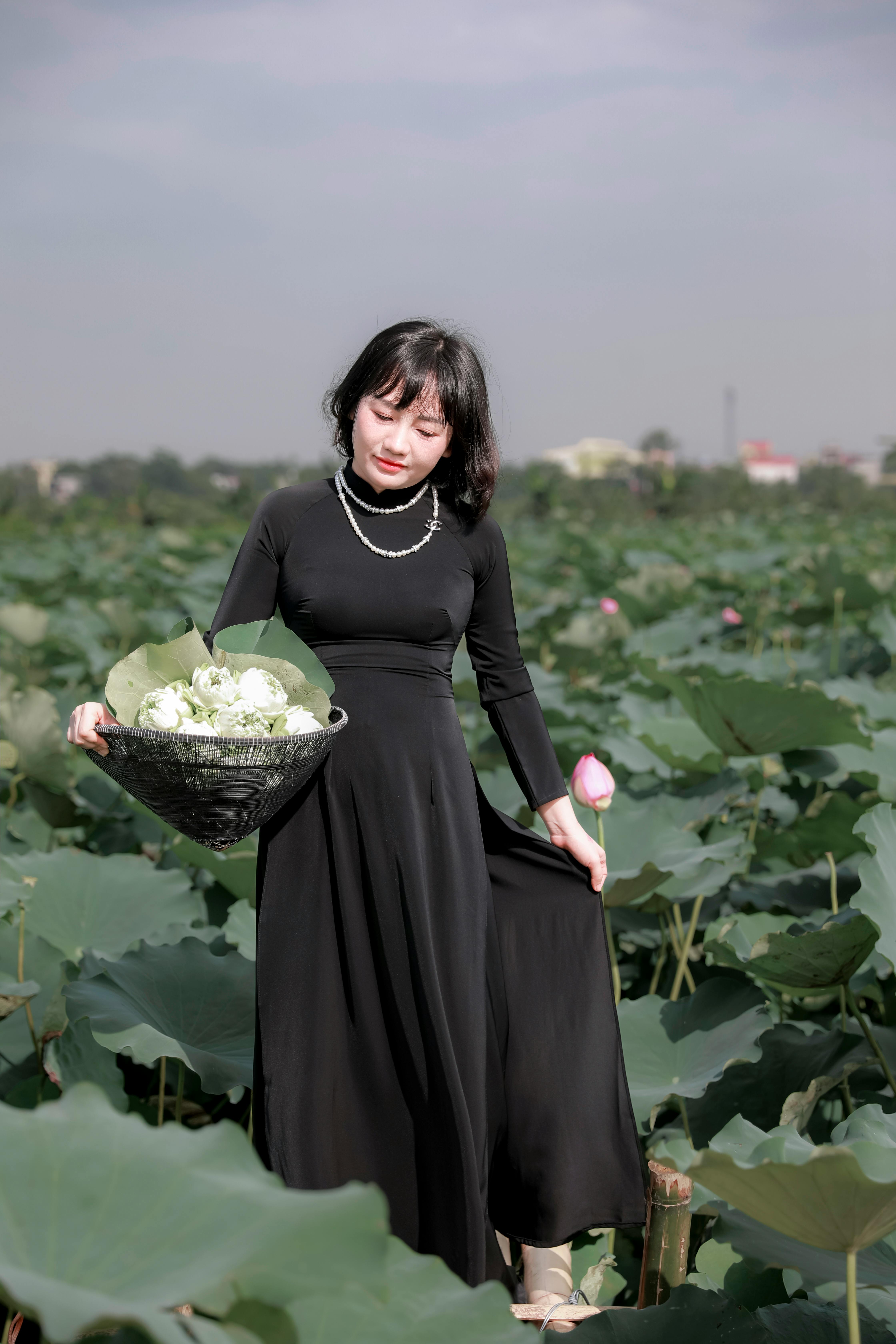 A young woman in a black Ao Dai holding flowers in a lush green field.