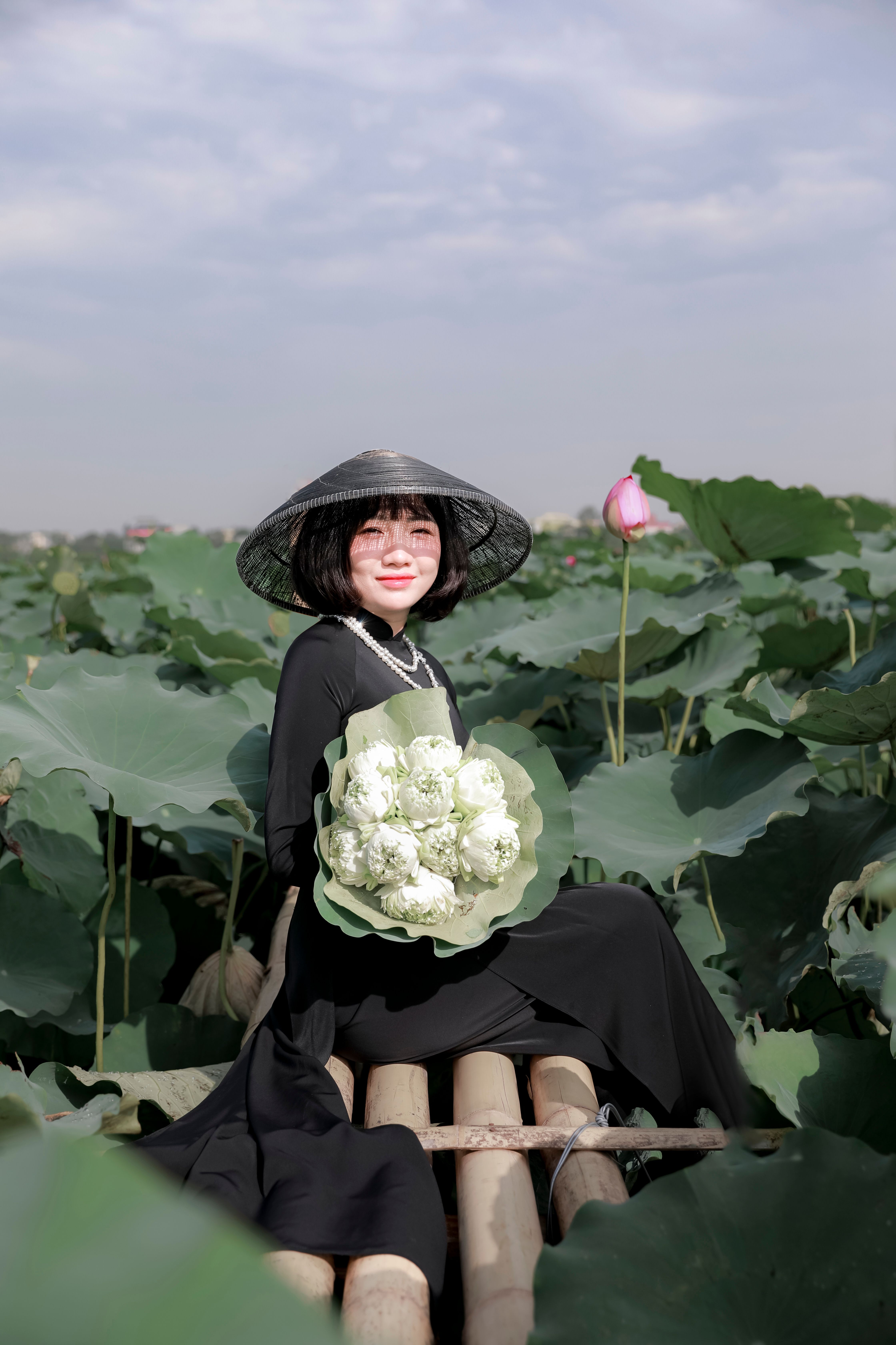 Asian woman in black dress holding flowers, posing amid green lotus leaves in a serene outdoor setting.
