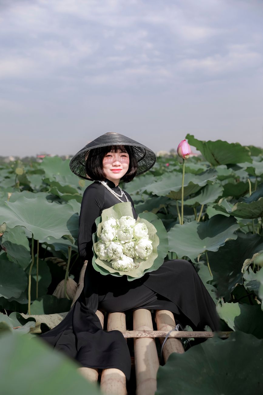Asian woman in black dress holding flowers, posing amid green lotus leaves in a serene outdoor setting.