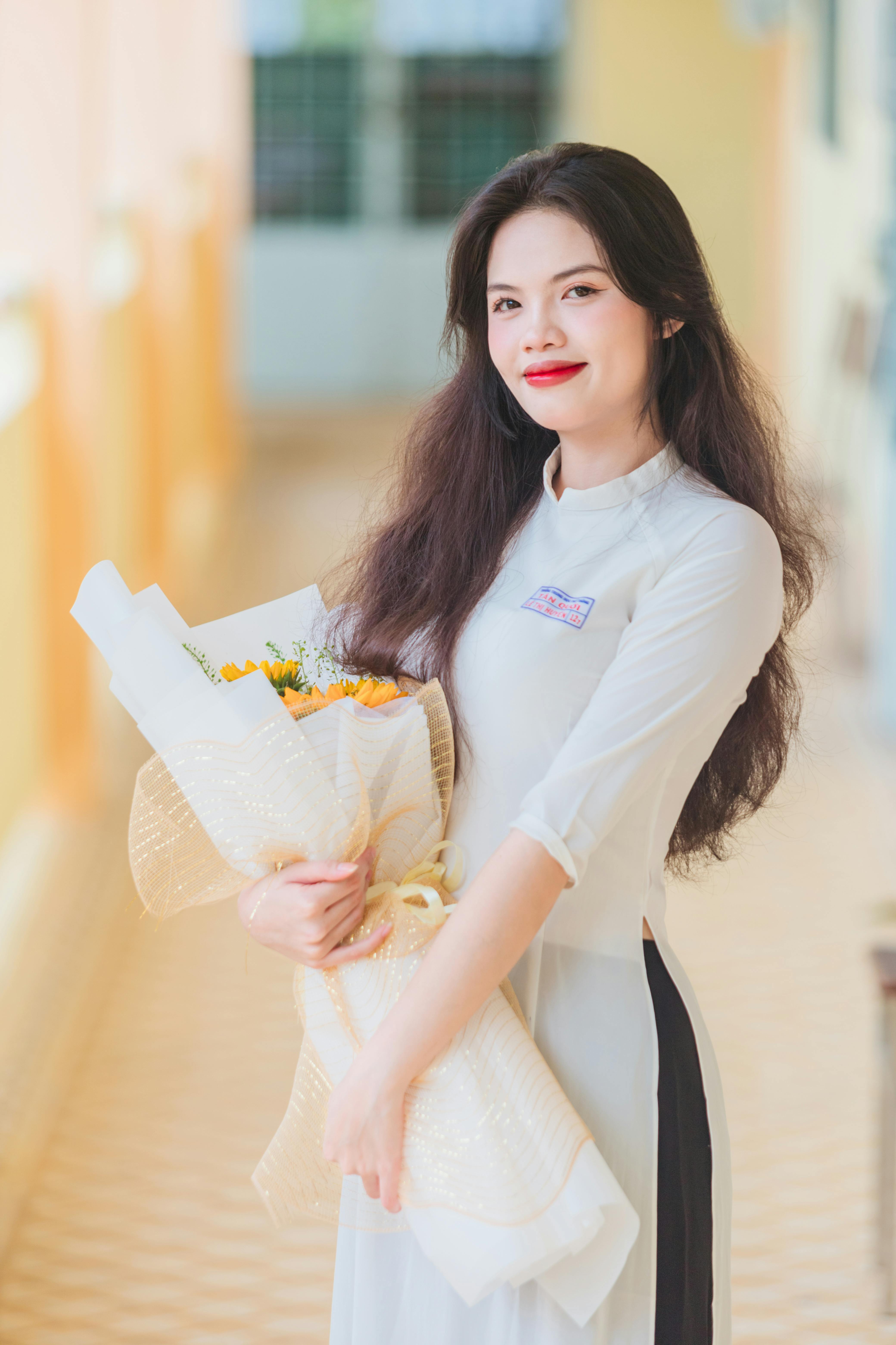 Asian woman wearing ao dai and holding a bouquet, smiling indoors.