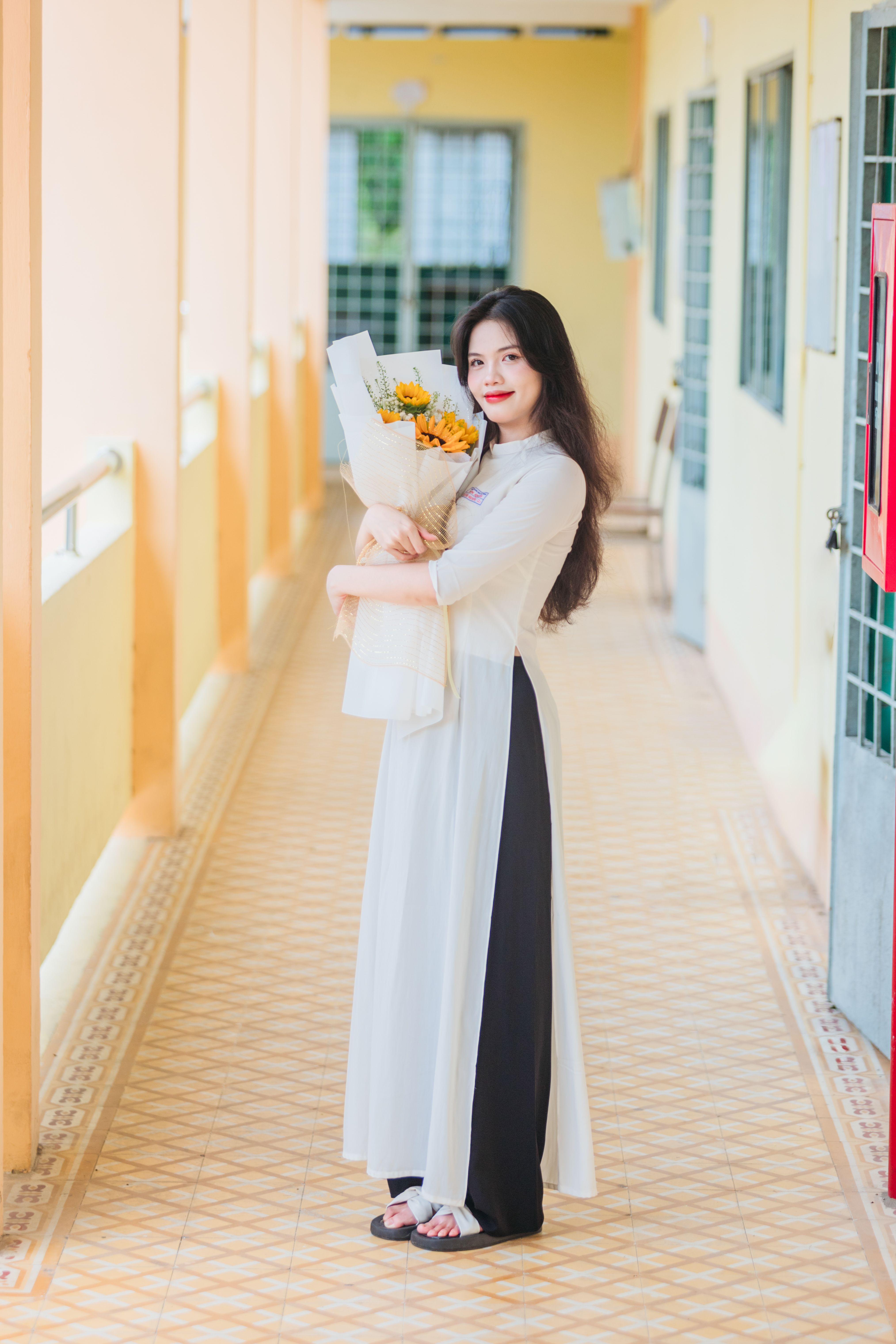 Young Vietnamese woman poses with a sunflower bouquet in a light-filled indoor hallway.