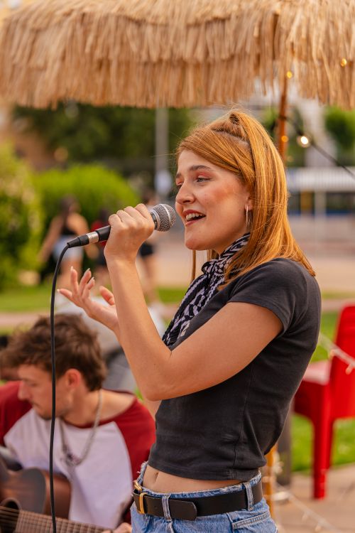 Free A young woman performs with a microphone at an outdoor concert, featuring a casual urban setting and acoustic guitar. Stock Photo