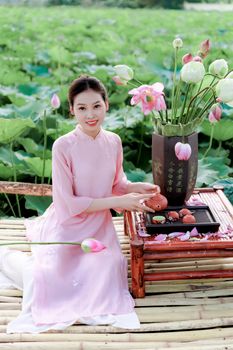 Young woman in pink ao dai, sitting peacefully by a lotus pond, enjoying tea.