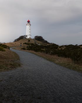 A solitary lighthouse stands on a grassy hill, guiding with its light under an overcast sky.