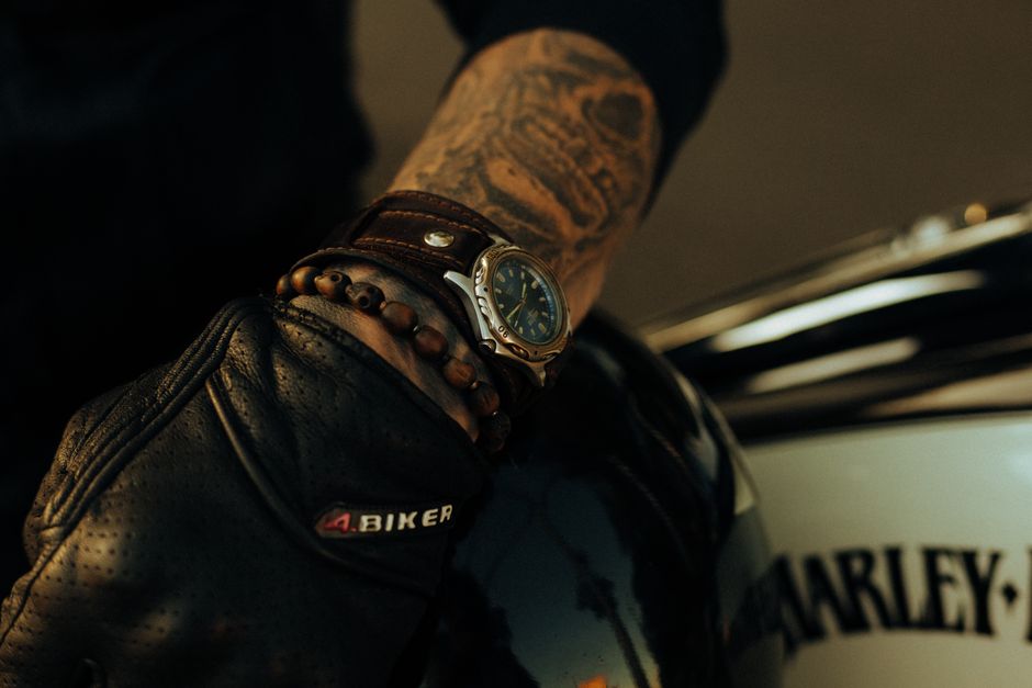 A masculine arm with tattoos wearing a watch and gloves, leaning on a motorcycle.