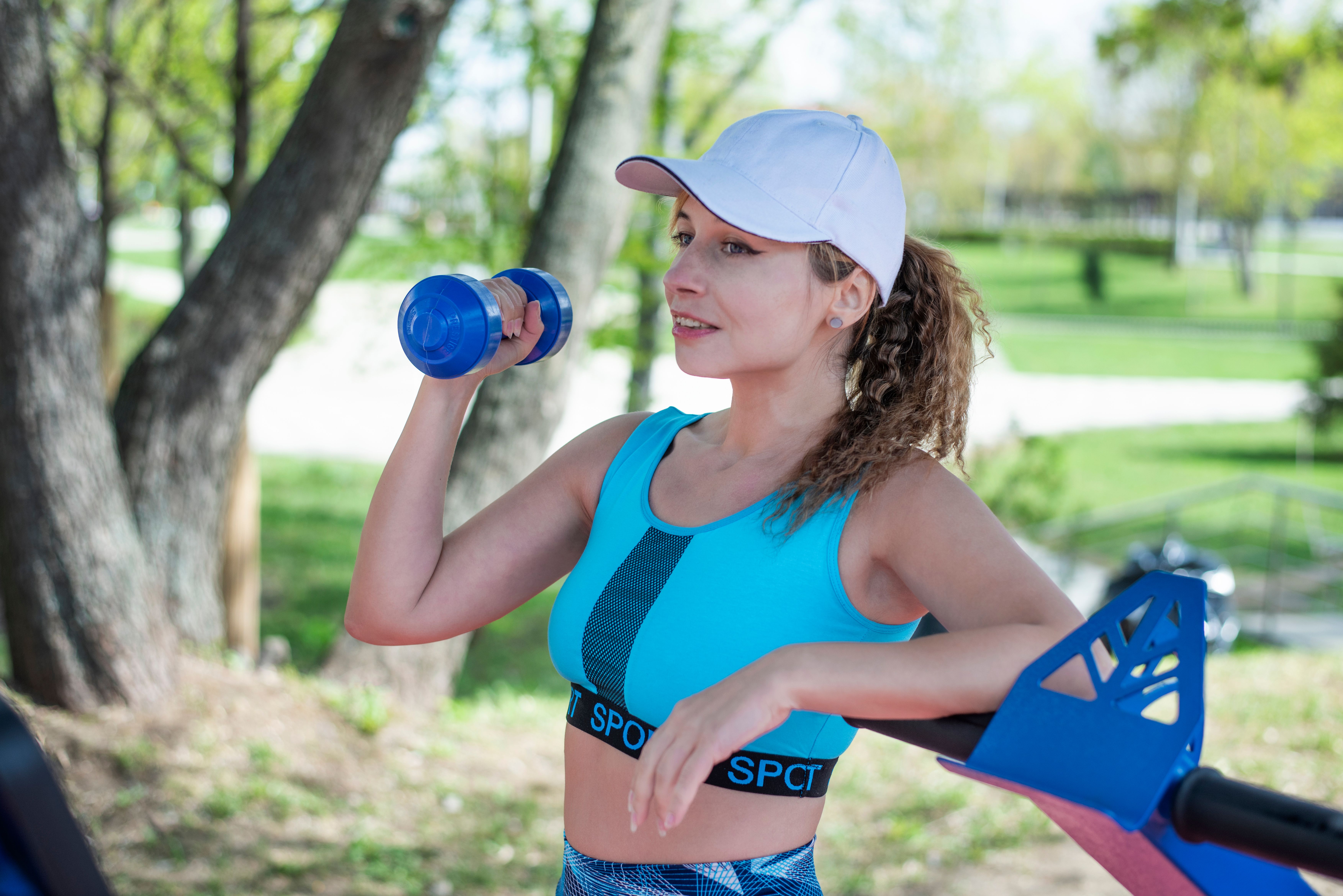 A woman in sportswear exercising with a dumbbell in a sunny park setting.