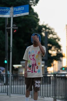 Fashionable man with blue hair poses on Paulista Avenue, showcasing urban streetwear style.