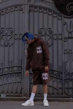 Stylish young man in bronze outfit and blue hair posing against ornate urban gate.