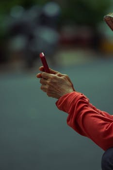 Close-up of hands using a smartphone in a red case outdoors. Focus on device, blurred background.