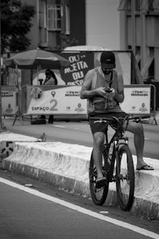 Black and white image of a young man sitting on a bicycle while using his phone in an urban environment.