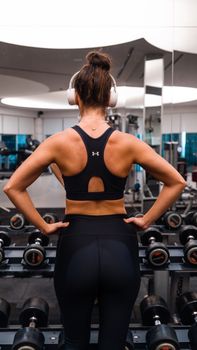 Fitness woman in gym with headphones standing by dumbbells. Back view.