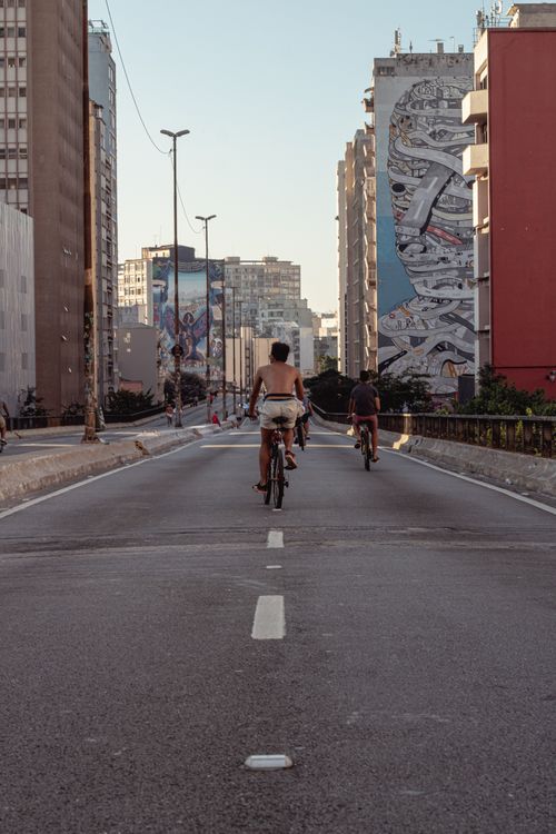 Free Cyclists ride through a city road bordered by tall buildings with vibrant graffiti art. Stock Photo