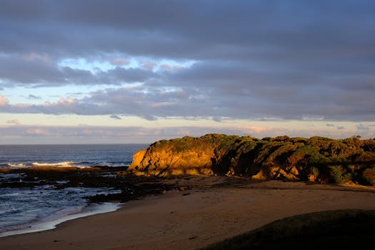 Serene beach scene at sunset with dramatic cliffs and ocean waves under a cloudy sky.