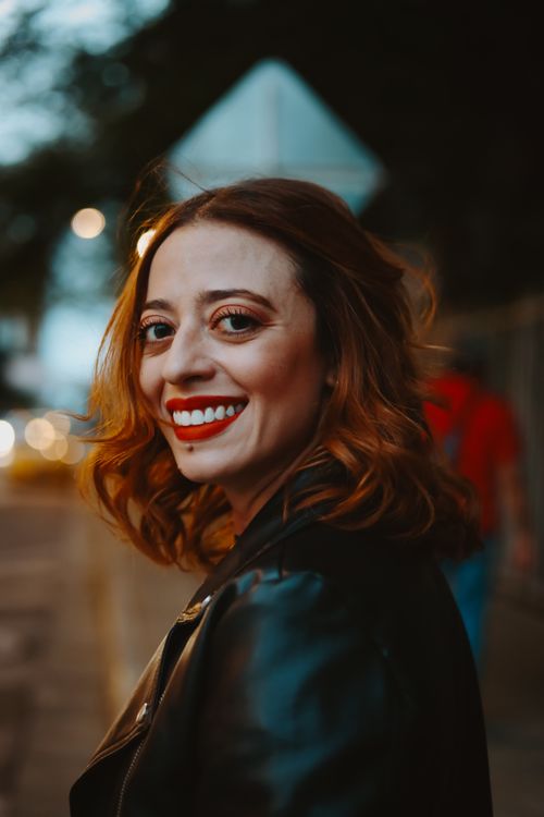 Free Portrait of a joyful redhead woman with red lipstick smiling outdoors in a leather jacket. Stock Photo