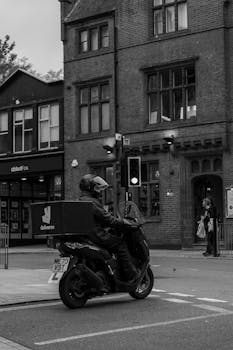 Black and white image of a delivery driver on a scooter waiting at a city intersection traffic light.