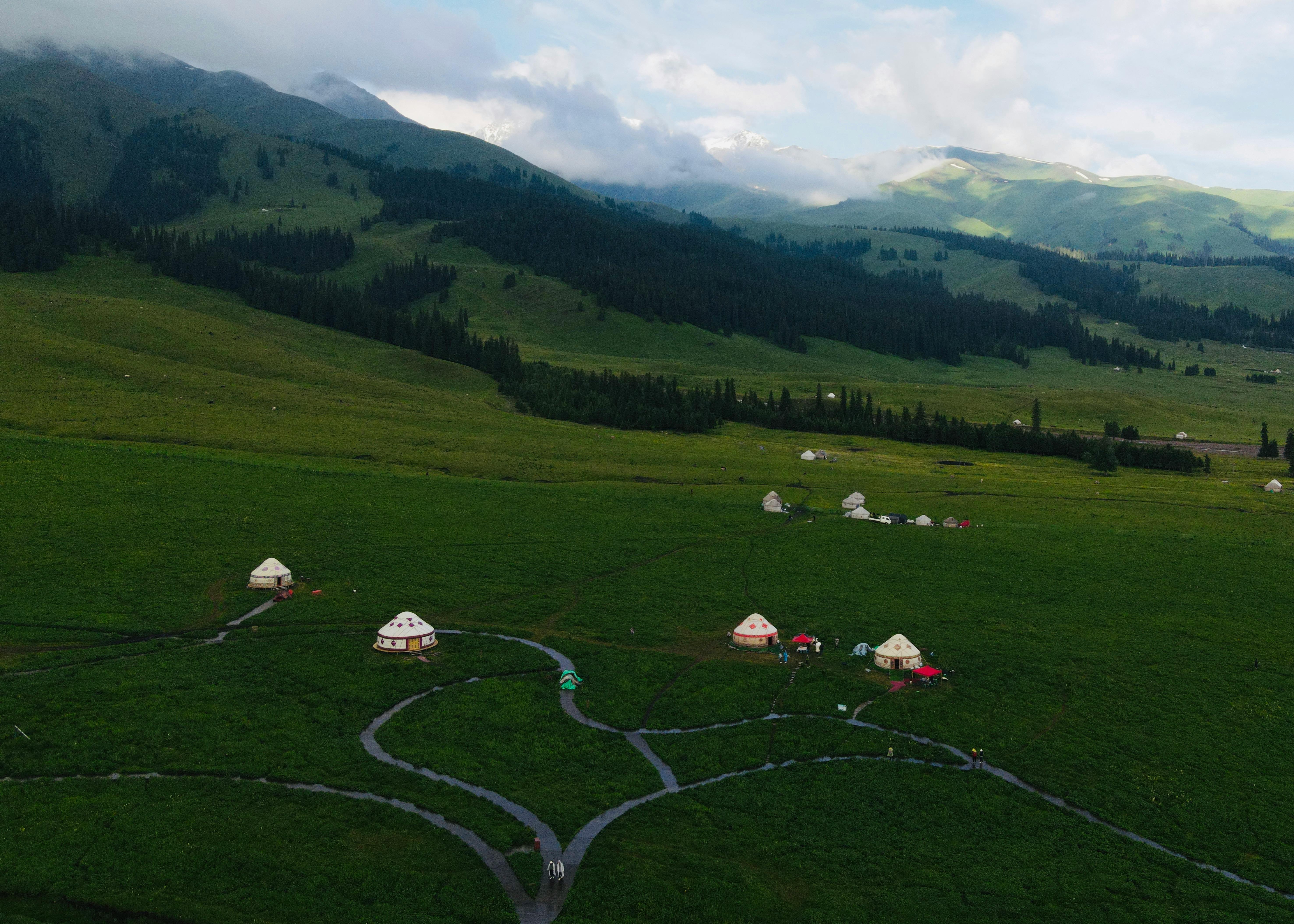 Explore the sprawling green hills and traditional yurts nestled in a tranquil countryside setting from above.
